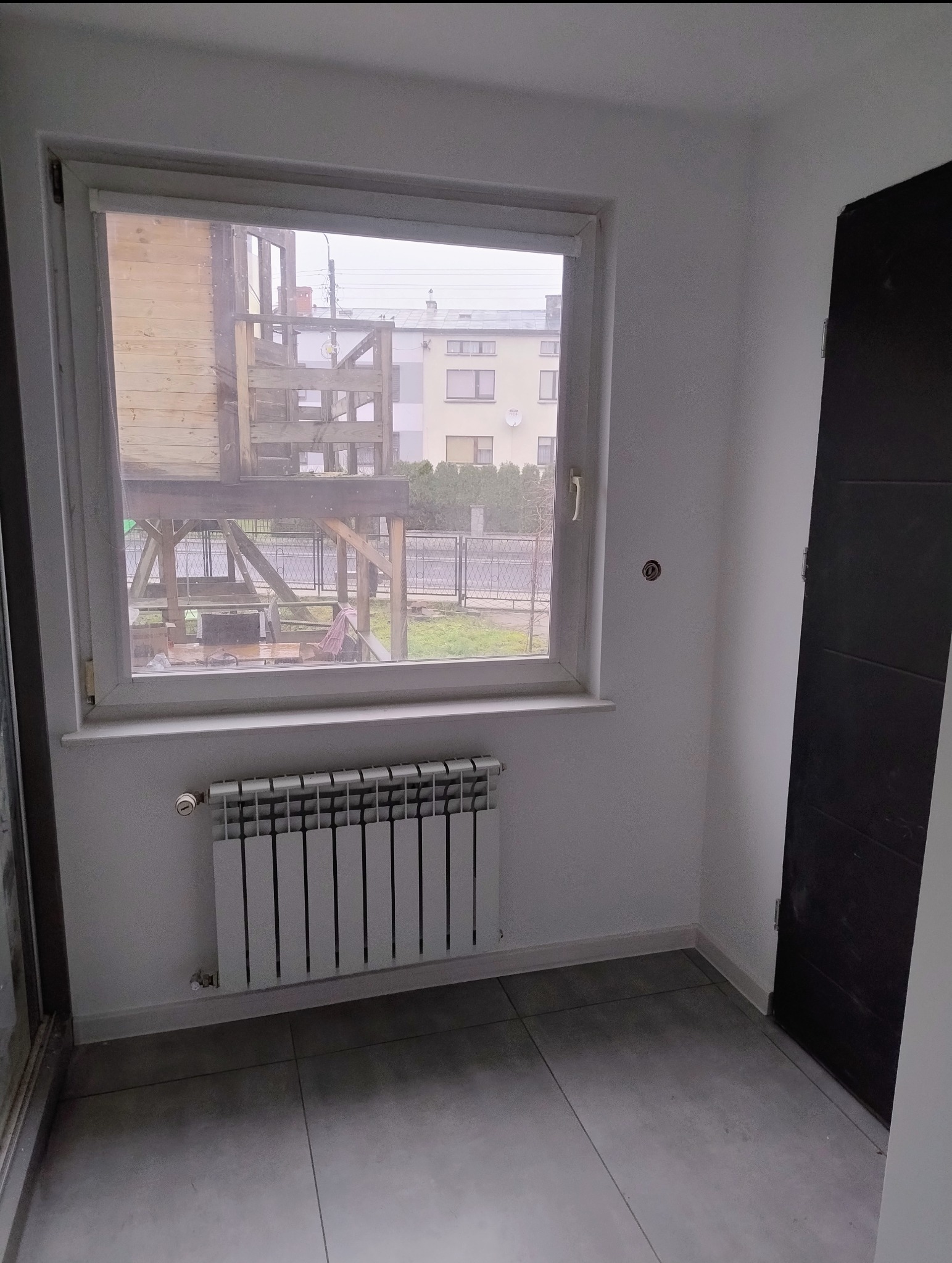 Wnętrze pokoju po remoncie: szare płytki podłogowe, białe ściany, grzejnik pod oknem z widokiem na plac zabaw i budynki. Ciemne drzwi po prawej stronie.