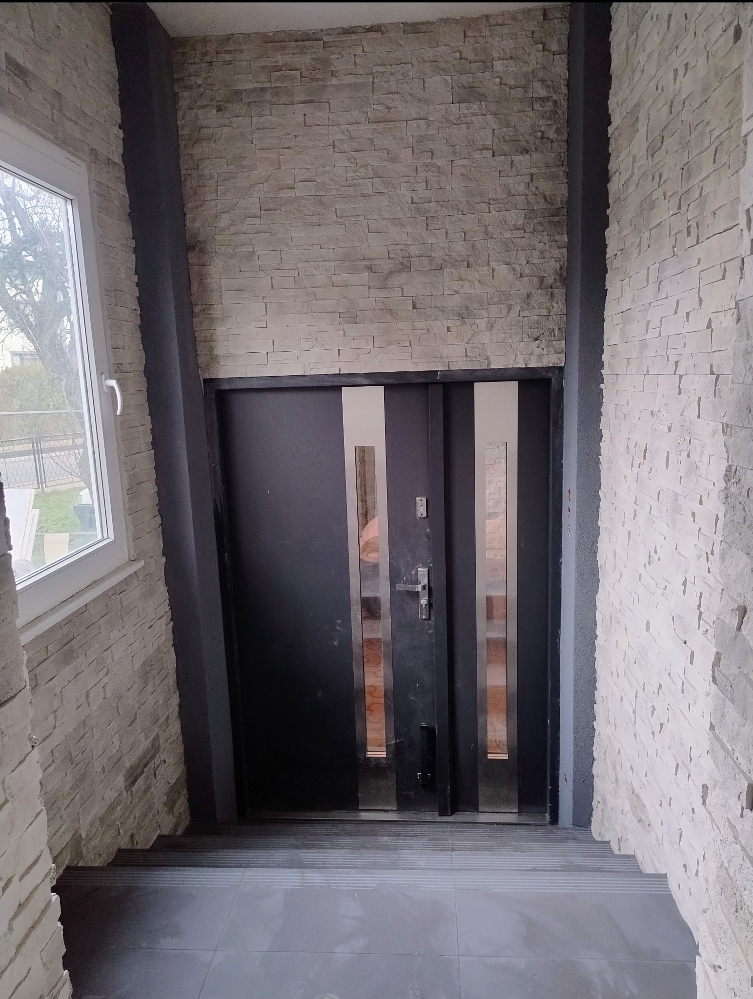 Wykończone wejście do domu z czarnymi drzwiami z przeszkleniami i kamienną okładziną ścian w odcieniach bieli i szarości. Widoczne schody z szarych płytek.