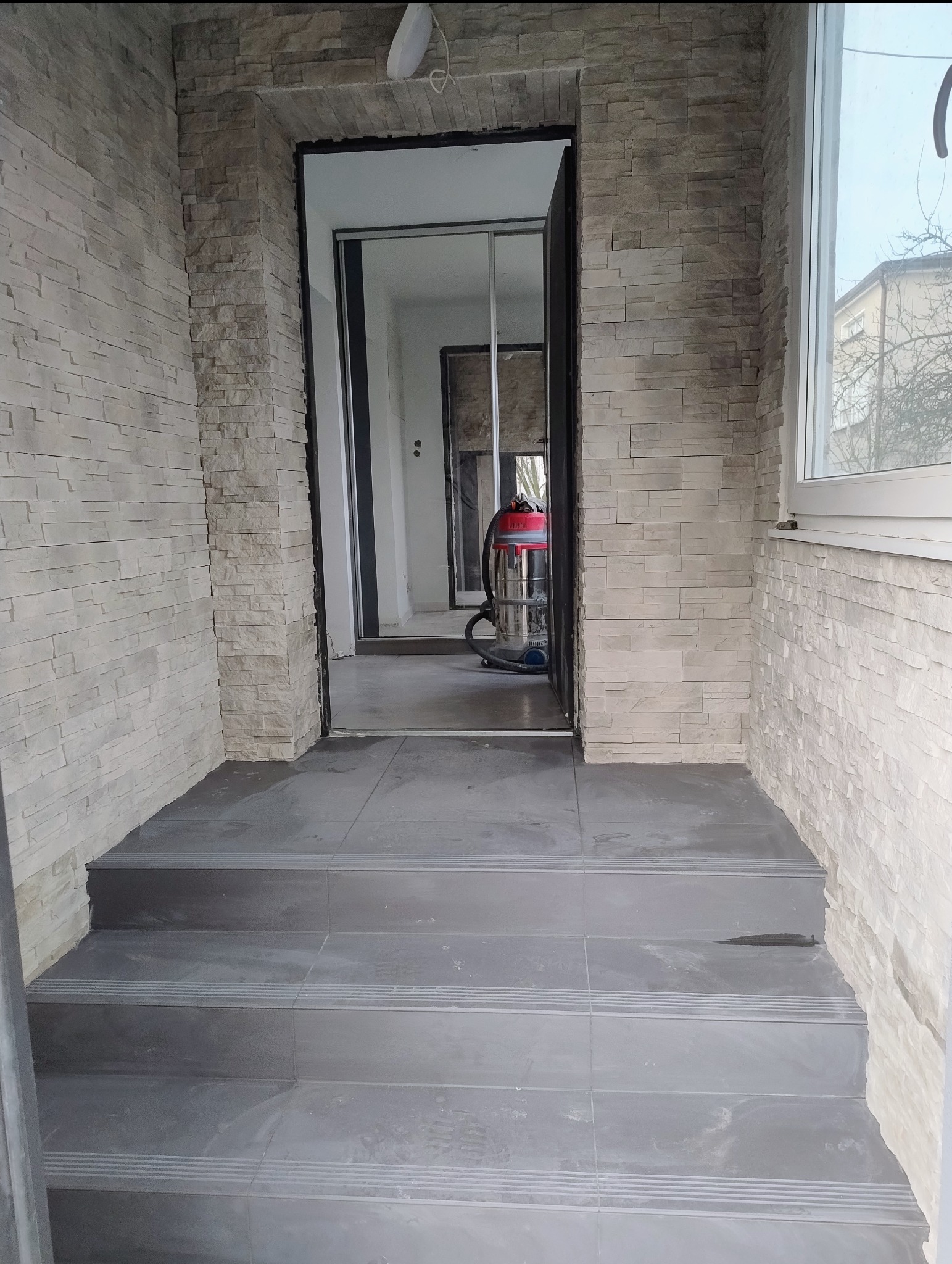 Wykończone schody wejściowe z szarych płytek, ściany wyłożone dekoracyjnym kamieniem w odcieniach beżu. W tle widoczny odkurzacz przemysłowy i przesuwne drzwi.