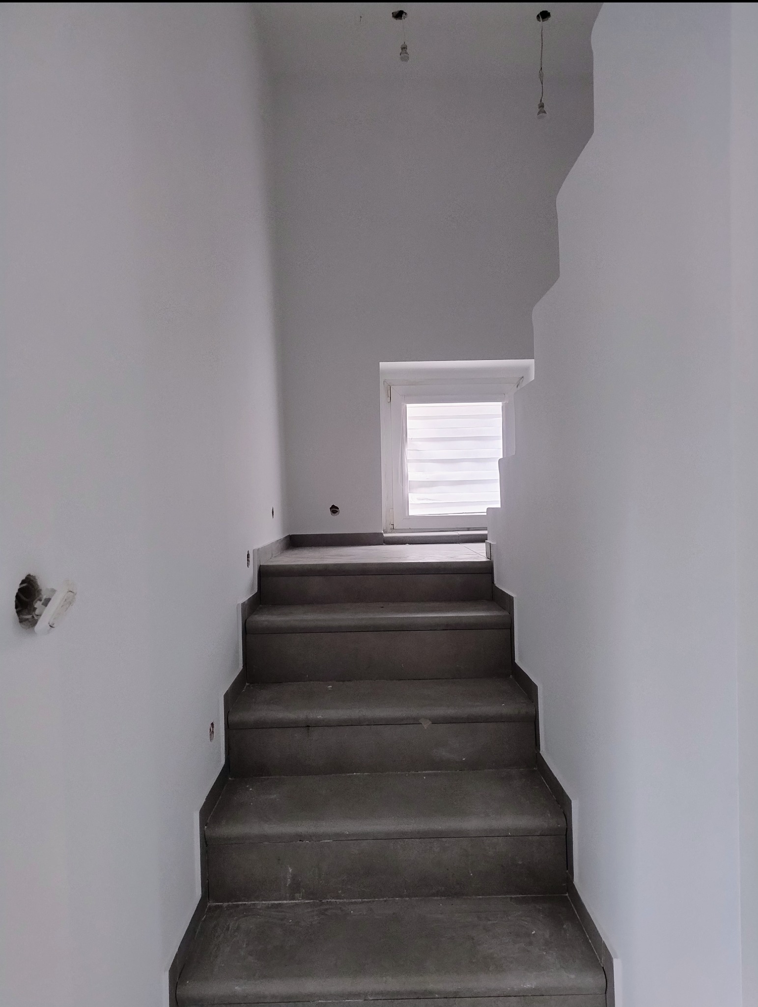 Schody z szarego kamienia prowadzące do okna z żaluzjami w białym, świeżo pomalowanym wnętrzu. Widoczne niedokończone punkty oświetleniowe.