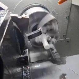 Precyzyjna obróbka CNC białego elementu na tokarce, w chłodzeniu emulsją. Wióry i płyn chłodzący widoczne w trakcie pracy. Dynamiczne ujęcie z bliska.