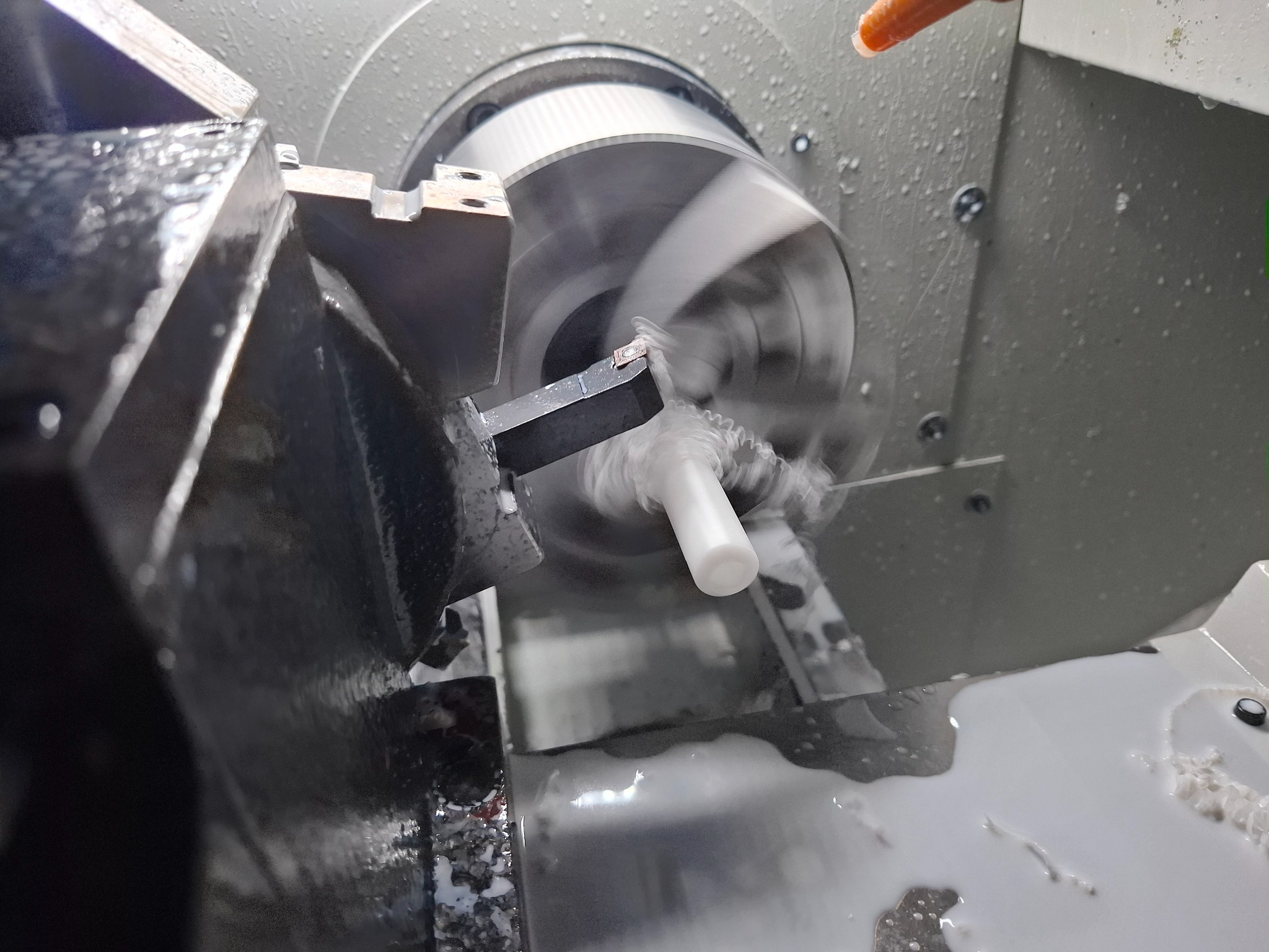 Precyzyjna obróbka CNC białego elementu na tokarce, w chłodzeniu emulsją. Wióry i płyn chłodzący widoczne w trakcie pracy. Dynamiczne ujęcie z bliska.