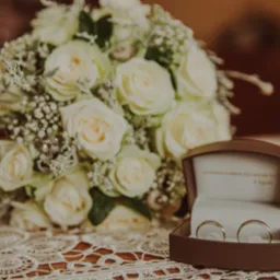 Bukiet białych róż i obrączki w brązowym pudełku na koronkowej serwetce. Subtelne, ciepłe oświetlenie, delikatne detale. Romantyczna fotografia ślubna.