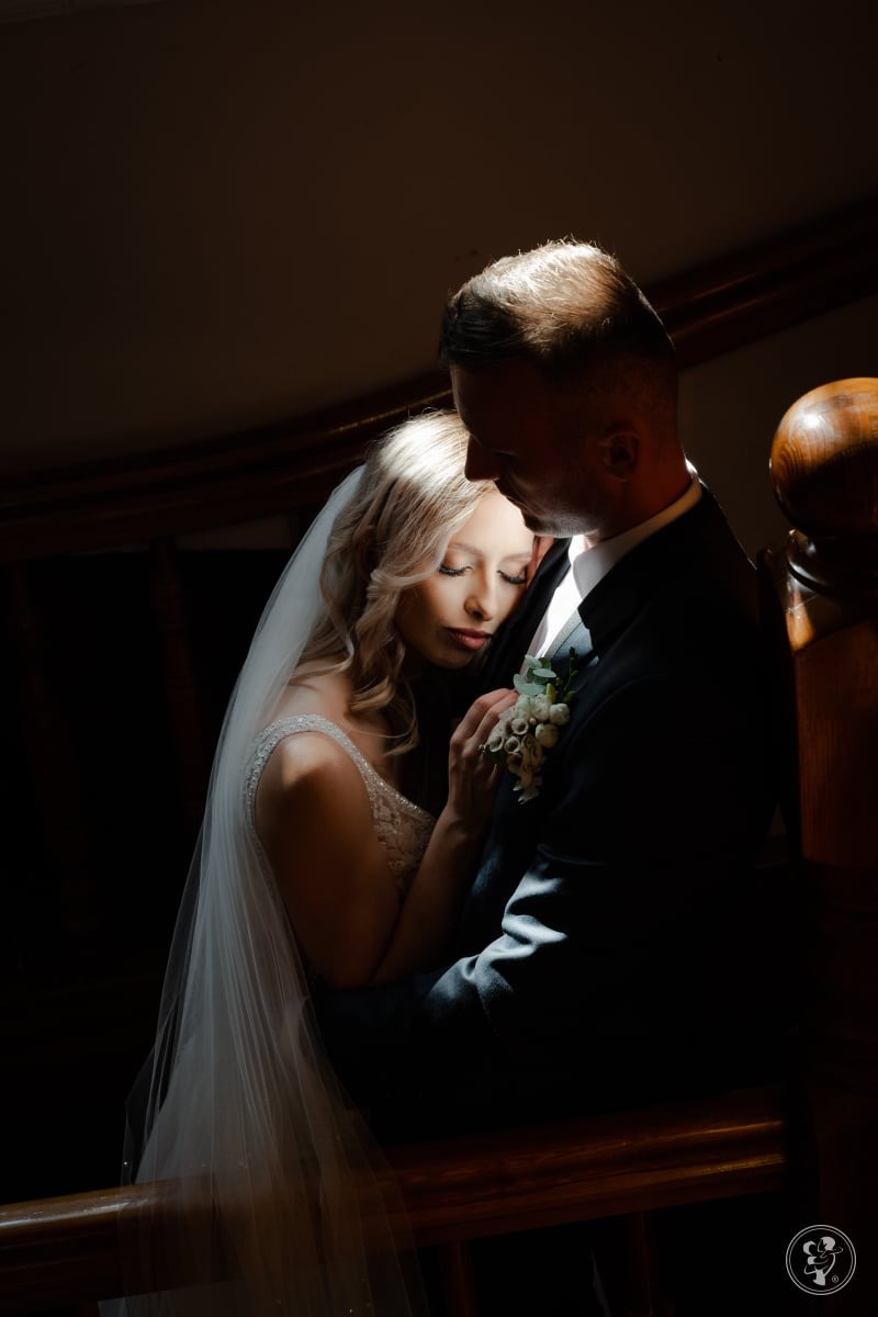 Zamyślona para młoda w objęciach, wpadające światło podkreśla emocje. Panna młoda w welonie, pan młody w ciemnym garniturze, delikatny bukiecik w butonierce.