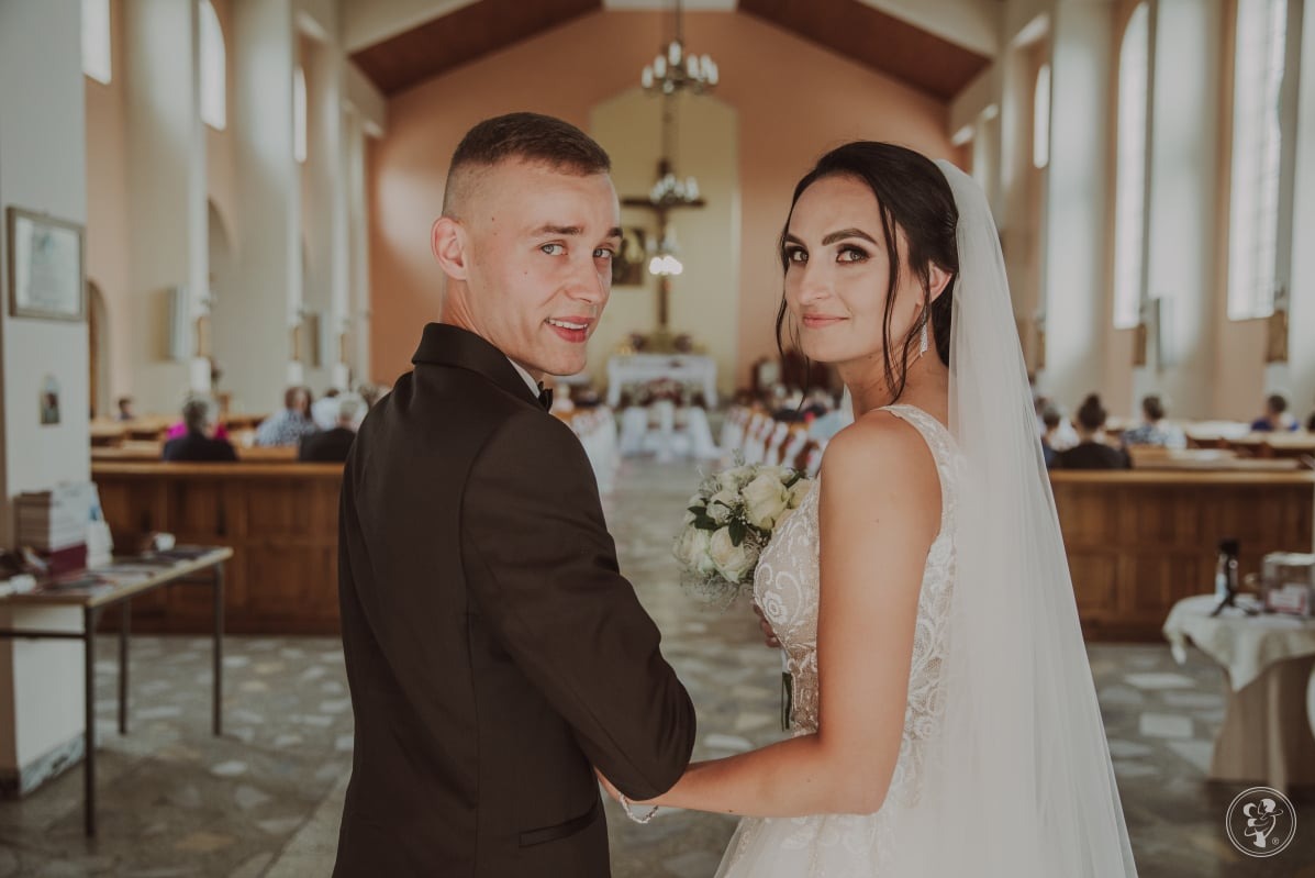 Para młoda, panna młoda w sukni z bukietem, pan młody w smokingu, oboje patrzą w obiektyw na tle wnętrza kościoła z wiernymi.