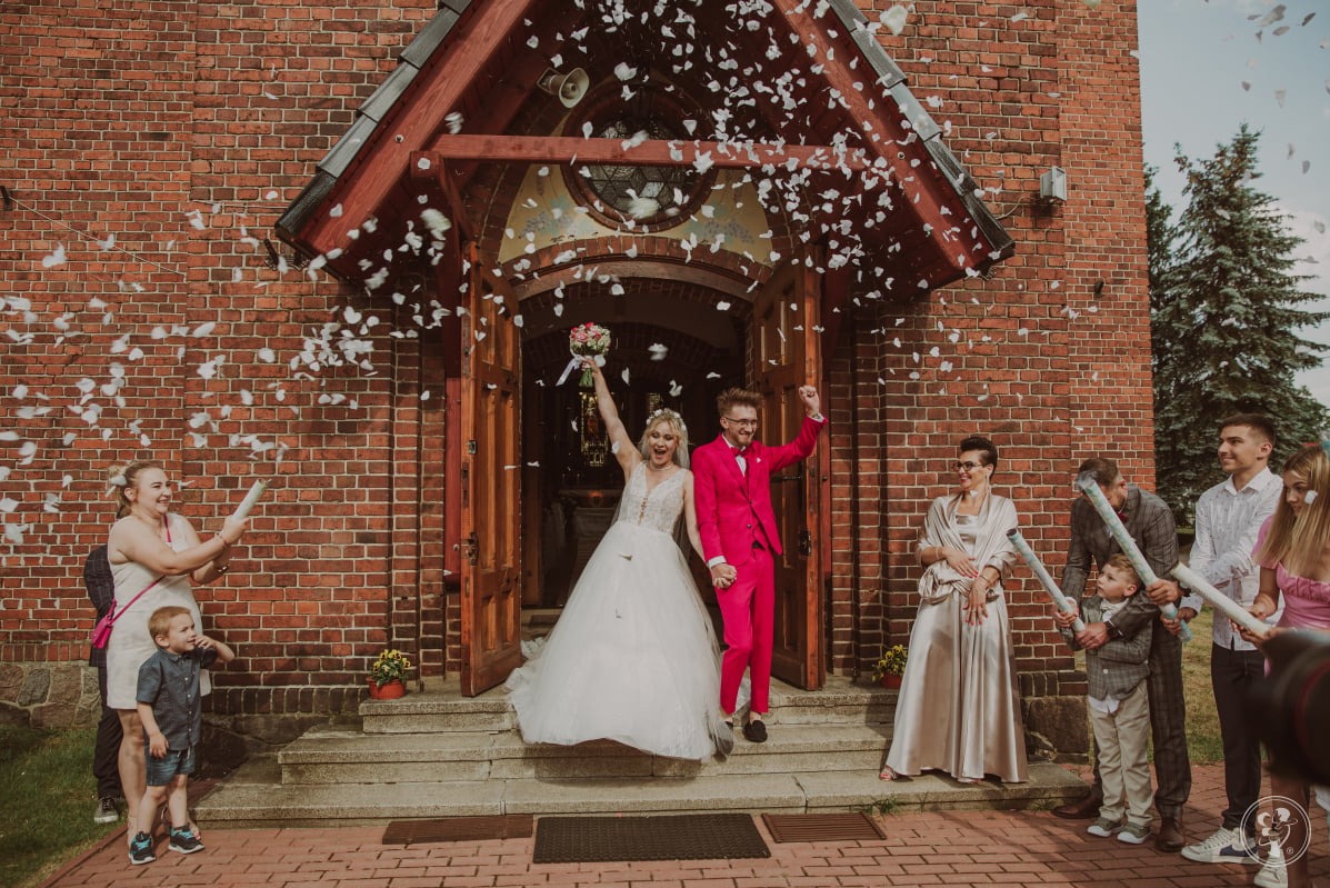 Para młoda w różowym garniturze i białej sukni wychodzi z kościoła, obsypywana konfetti, z uśmiechami na twarzach. Goście weselni w tle.