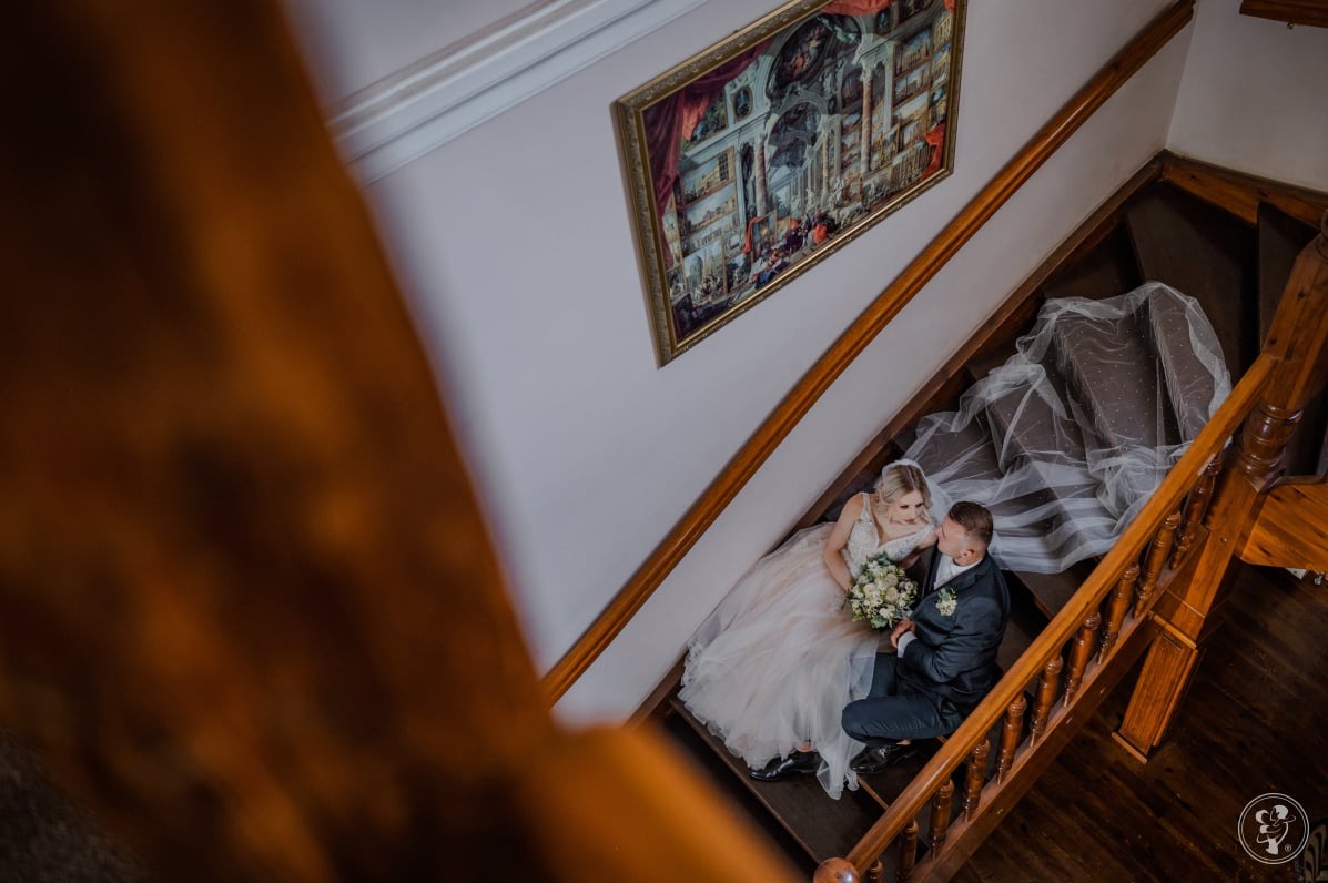 Para młoda siedzi na drewnianych schodach, panna młoda w sukni z bukietem, pan młody w garniturze. Ujęcie z góry, w tle obraz na ścianie.