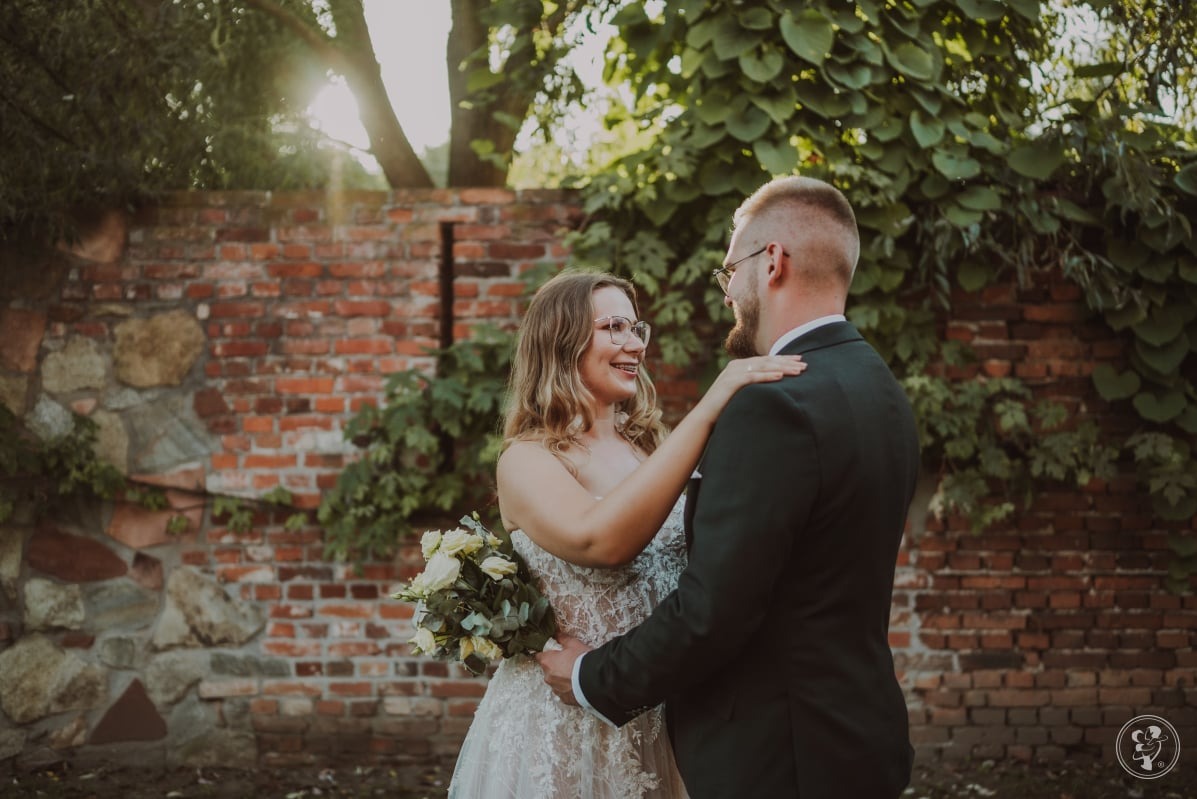 Uśmiechnięta panna młoda w okularach i koronkowej sukni obejmuje pana młodego w ciemnym garniturze na tle ceglanego muru z bluszczem w słoneczny dzień.