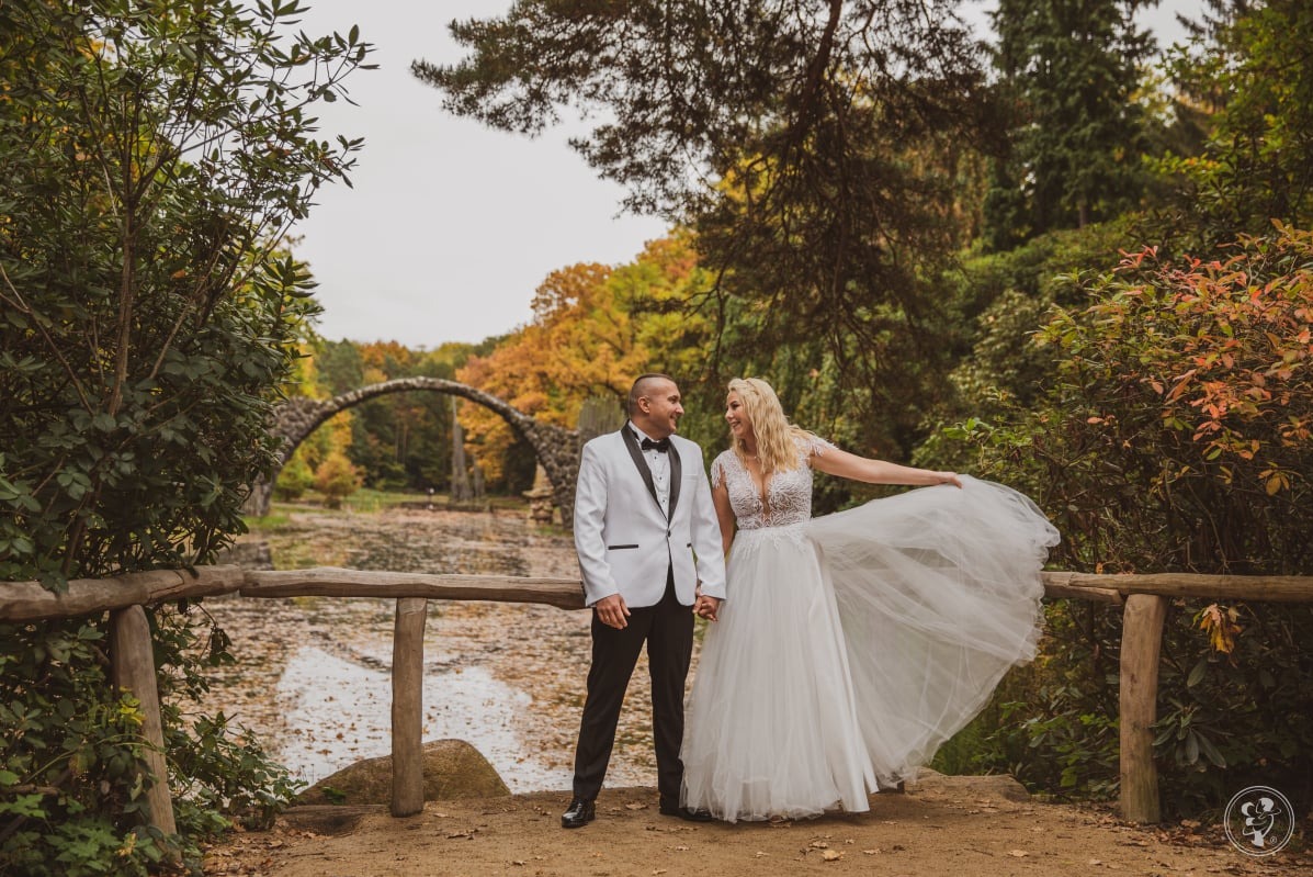 Para młoda trzymająca się za ręce na tle Rakotzbrücke w Kromlau. Panna młoda w koronkowej sukni, pan młody w białym smokingu. Jesienna sceneria parku.