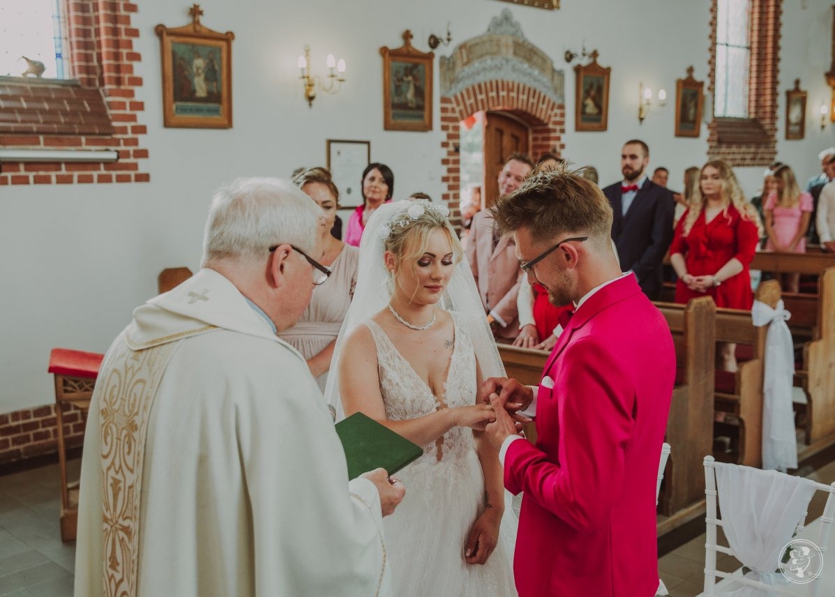 Para młoda wymienia się obrączkami podczas ceremonii ślubnej w kościele. Pan młody w różowym garniturze. W tle goście i ksiądz.