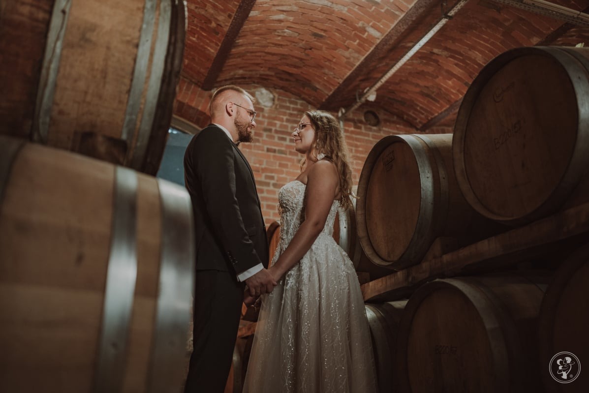 Para młoda, on w czarnym garniturze, ona w białej sukni, trzymają się za ręce w piwnicy z beczkami, ceglany sufit, ciepłe oświetlenie, spojrzenie pełne miłości.