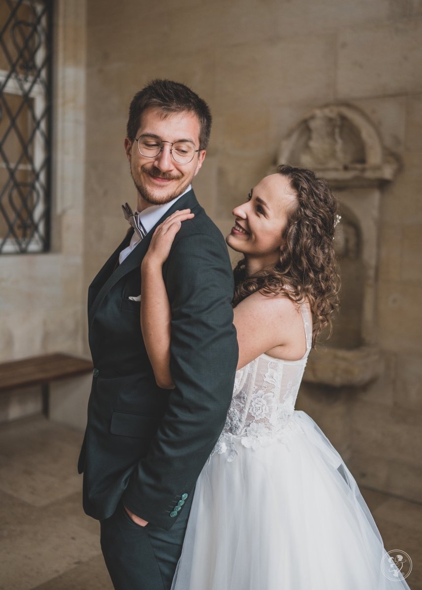 Uśmiechnięta panna młoda obejmuje eleganckiego pana młodego w ciemnozielonym garniturze i muszce, na tle kamiennej ściany z dekoracyjną wnęką i ławką.