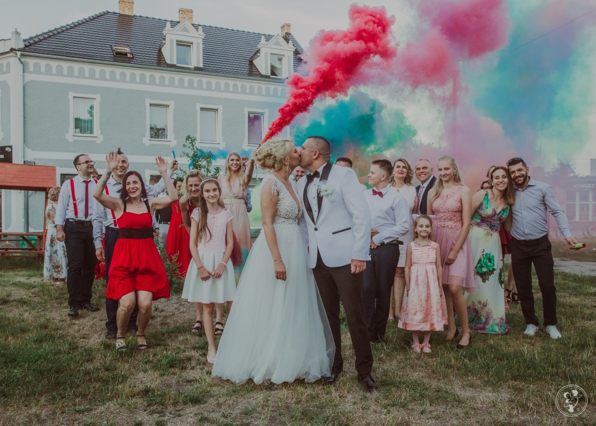 Para młoda całuje się na tle gości i kolorowych dymów podczas wesela. Uśmiechnięci ludzie, radosna atmosfera, budynek w tle.