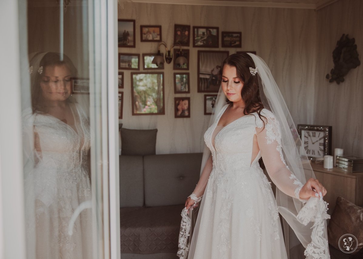 Panna młoda w koronkowej sukni ślubnej z welonem, w jasnym wnętrzu z ramkami na ścianie. Widoczne odbicie w szklanych drzwiach.