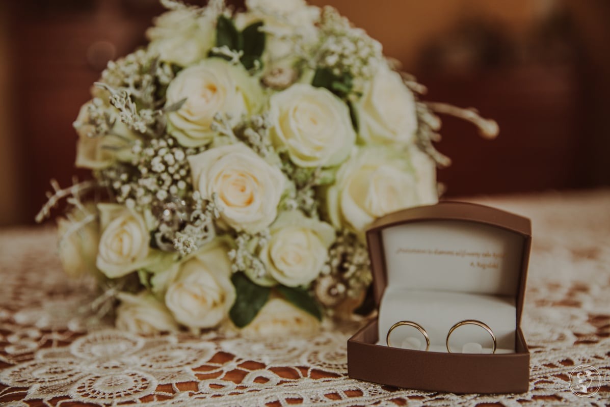 Bukiet białych róż i obrączki w brązowym pudełku na koronkowej serwetce. Subtelne, ciepłe oświetlenie, delikatne detale. Romantyczna fotografia ślubna.