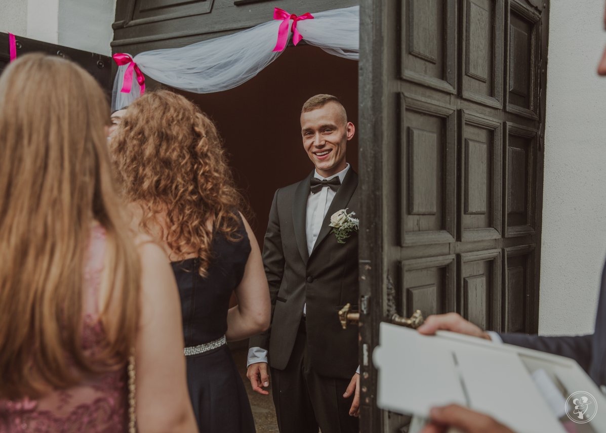 Uśmiechnięty pan młody w czarnym smokingu i muszce, czeka w drzwiach udekorowanych różowymi wstążkami, na gości weselnych.