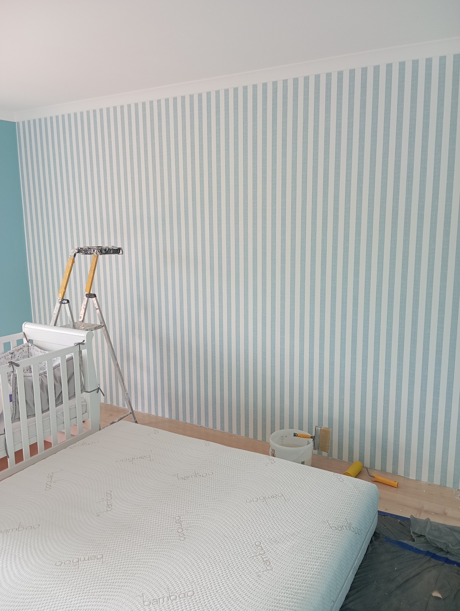 Ściana w pokoju dziecięcym po malowaniu w biało-błękitne pasy. Widoczny drabina malarska, wałek i wiadro z farbą. Częściowo widoczne łóżeczko dziecięce.
