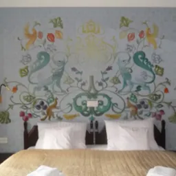 Sypialnia z łóżkiem i artystycznym malowaniem ściany z motywami zwierzęcymi i roślinnymi w stylu fantasy, oświetlona złotymi lampkami.