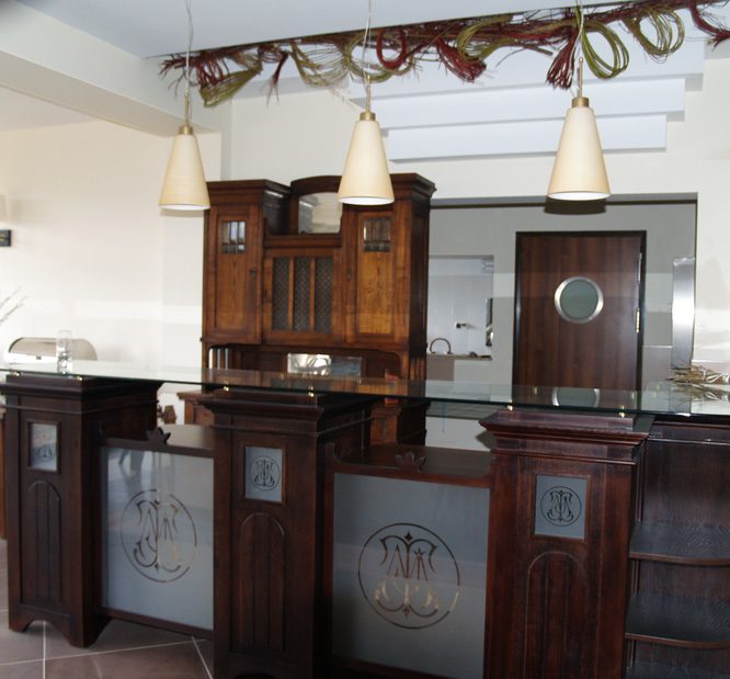 Recepcja w stylu retro z drewnianą zabudową, szklanym blatem i trzema stożkowymi lampami wiszącymi. Dekoracja z gałązek na suficie.