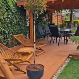 Zadaszony taras z drewna z leżakami, stołem i krzesłami. Widoczne rośliny w donicach i trawnik. Pergola z drewnianą konstrukcją.