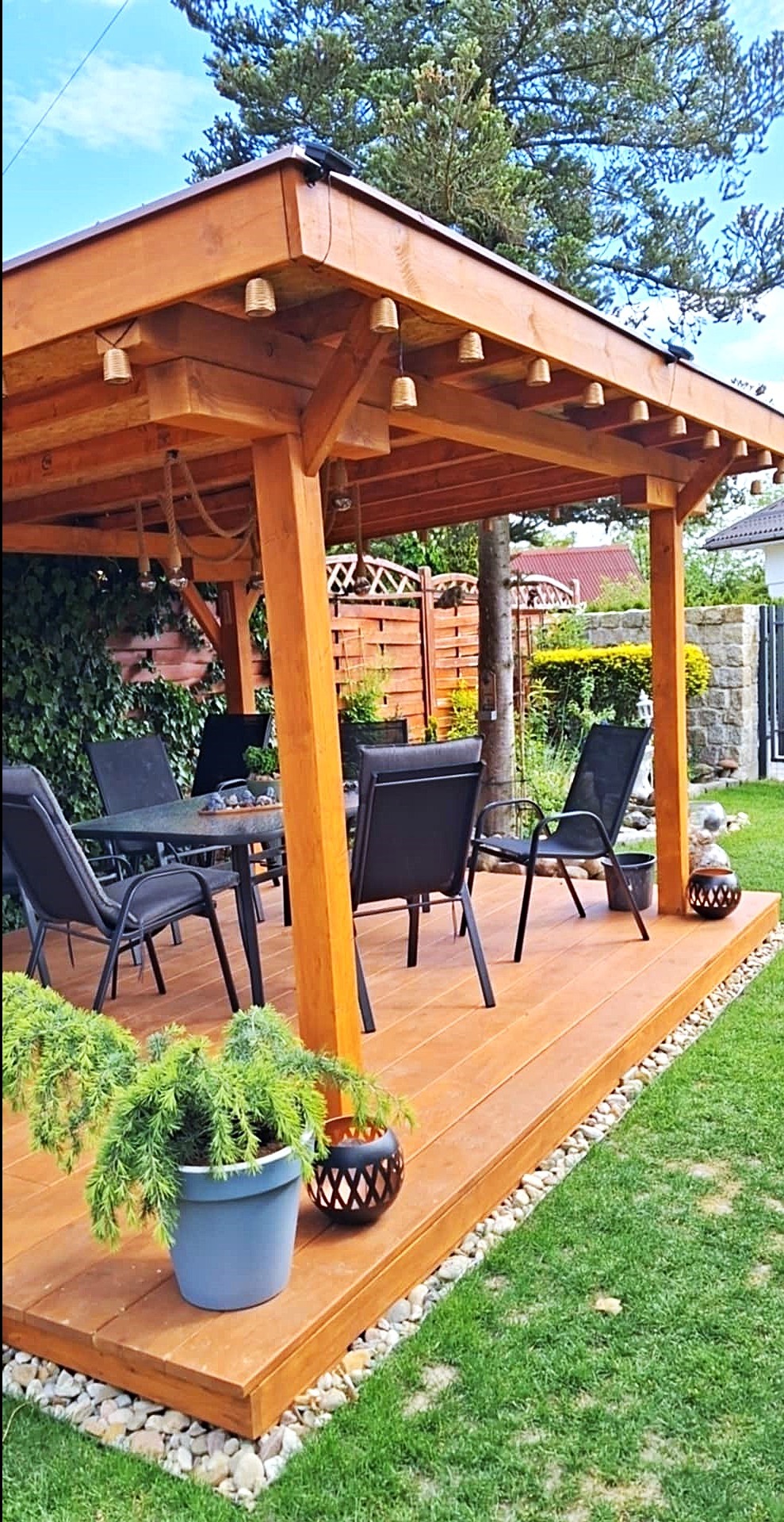 Zadaszony taras z drewna z oświetleniem w stylu boho, otoczony zielenią. Meble ogrodowe, dekoracyjne donice i kamienny obrzeże dodają uroku.