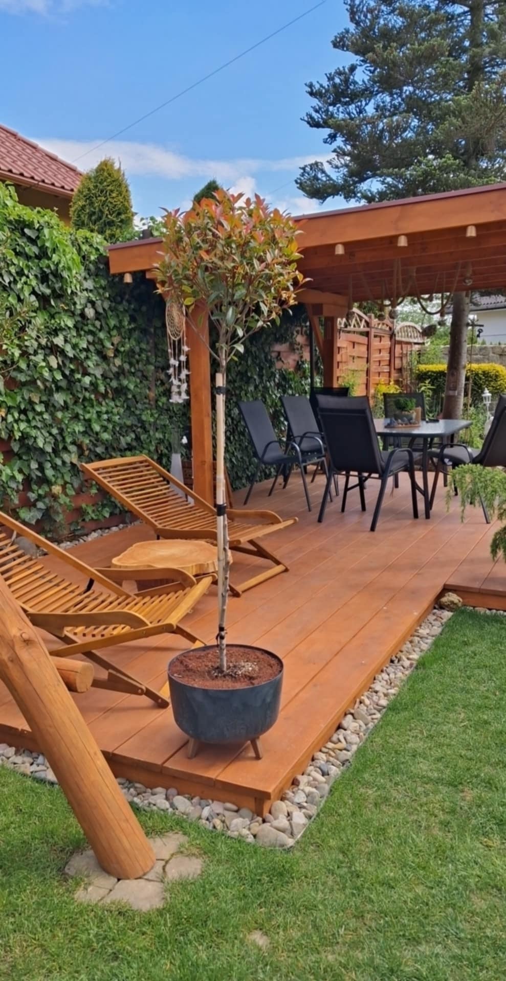 Zadaszony taras z drewna z leżakami, stołem i krzesłami. Widoczne rośliny w donicach i trawnik. Pergola z drewnianą konstrukcją.