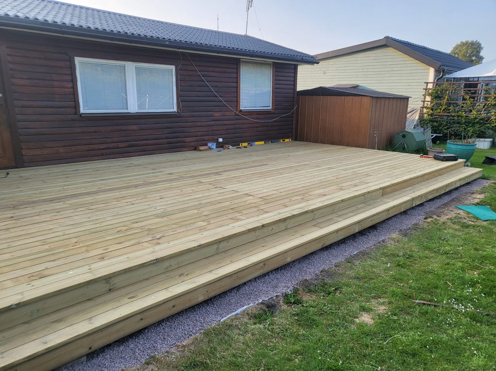 Nowy taras z drewna ze schodami, przylegający do brązowego domu z białymi ramami okiennymi. Na tarasie poziomica i inne narzędzia, obok trawnik.