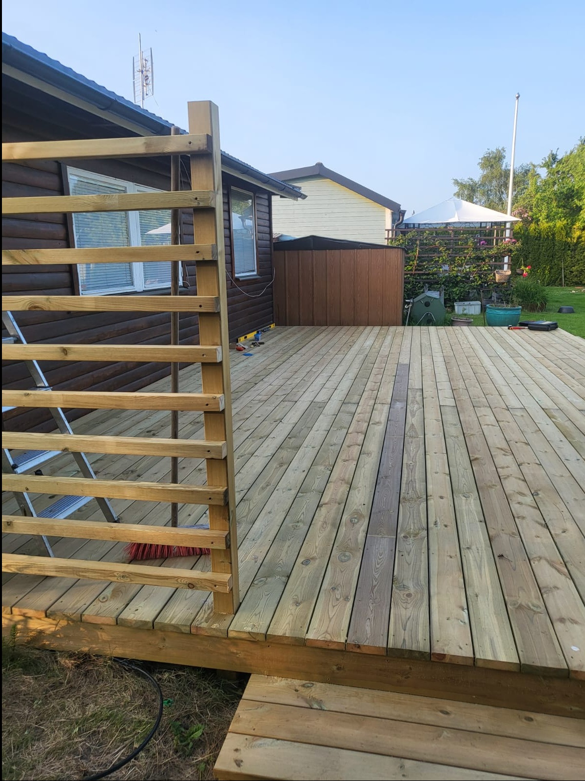 Drewniany taras z surowego drewna w ogrodzie, częściowo zadaszony, z widoczną konstrukcją ażurową i drabinką. W tle zabudowa ogrodowa i zieleń.