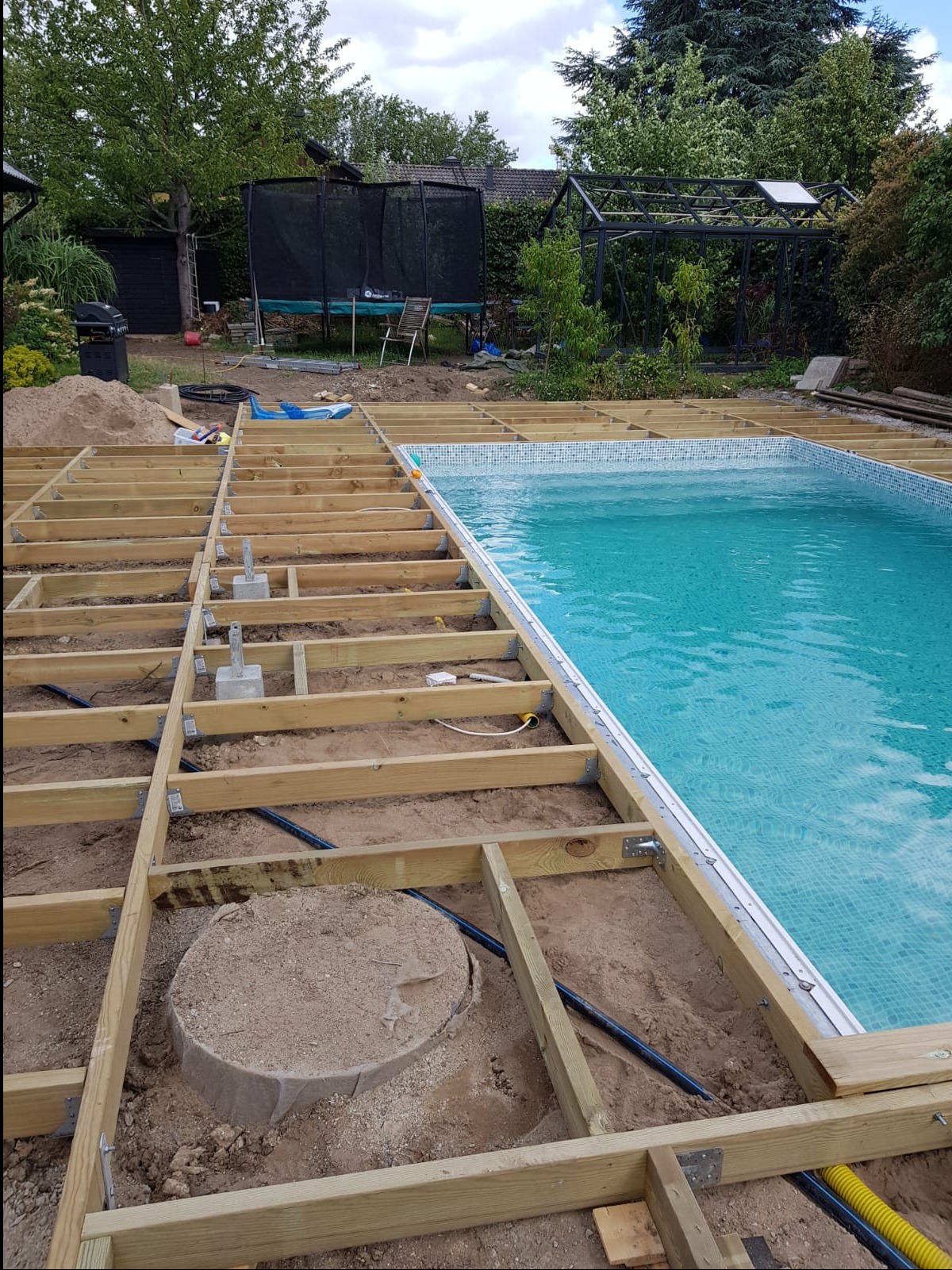 Konstrukcja pod taras przy basenie, z widocznymi elementami drewnianymi i betonowymi wspornikami, w tle ogród z trampoliną i szklarnią.