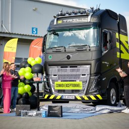 Goodbrand.ing - Uroczyste odsłonięcie czarnej ciężarówki marki Volvo z żółtymi akcentami, logiem 'Trill', balonami i osobami klaszczącymi w tle, przed budynkiem magazynowym.