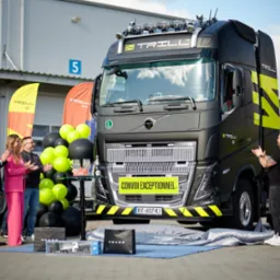 Uroczyste odsłonięcie czarnej ciężarówki marki Volvo z żółtymi akcentami, logiem 'Trill', balonami i osobami klaszczącymi w tle, przed budynkiem magazynowym.