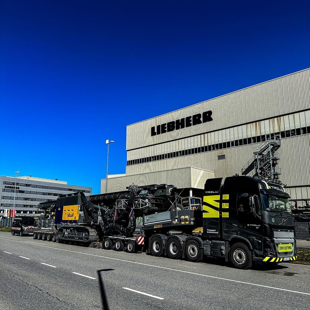 Czarna ciężarówka marki Volvo przewozi na lawecie maszynę budowlaną firmy Keller. W tle budynek z napisem Liebherr i błękitne niebo.