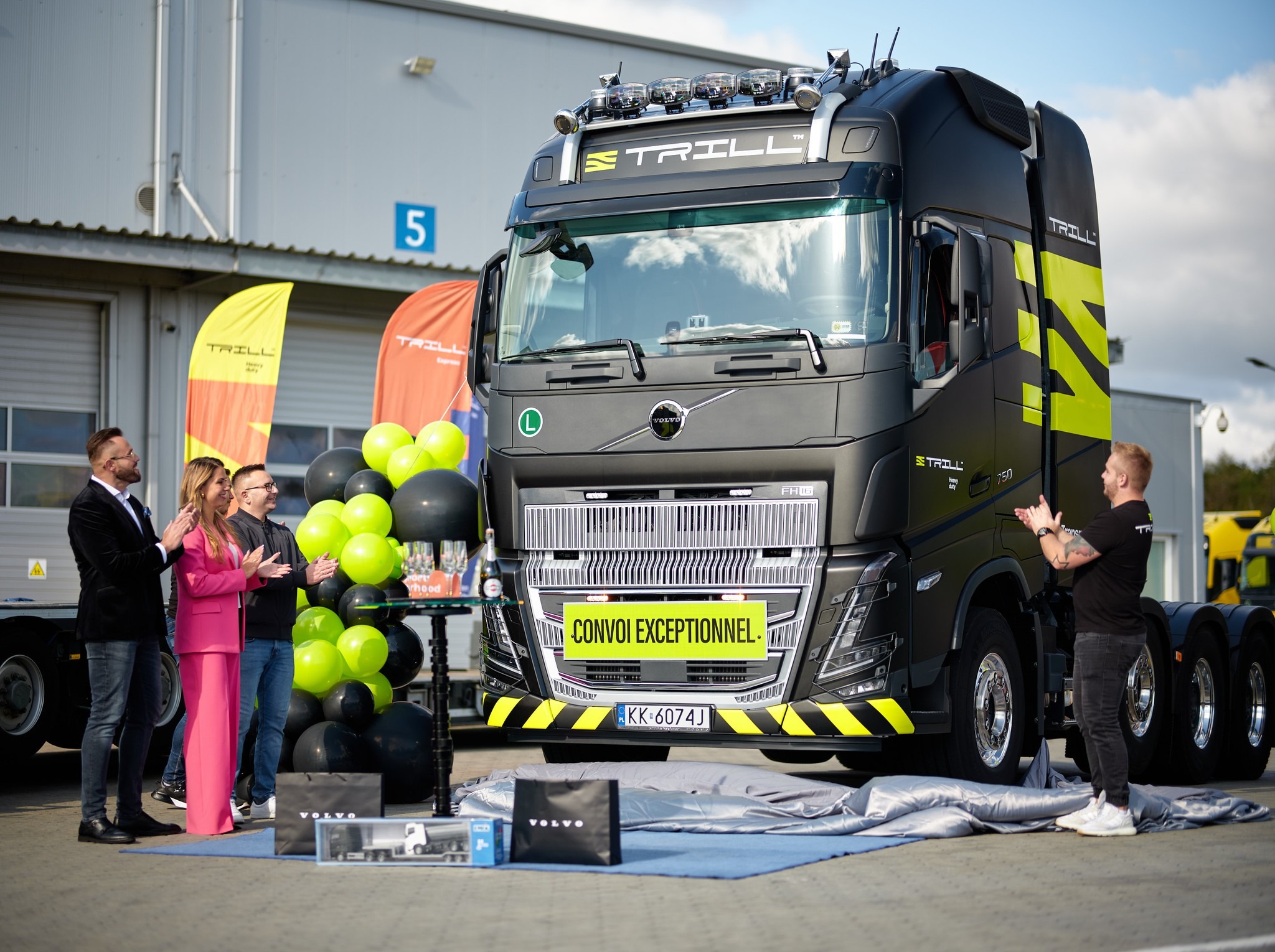 Uroczyste odsłonięcie czarnej ciężarówki marki Volvo z żółtymi akcentami, logiem 'Trill', balonami i osobami klaszczącymi w tle, przed budynkiem magazynowym.