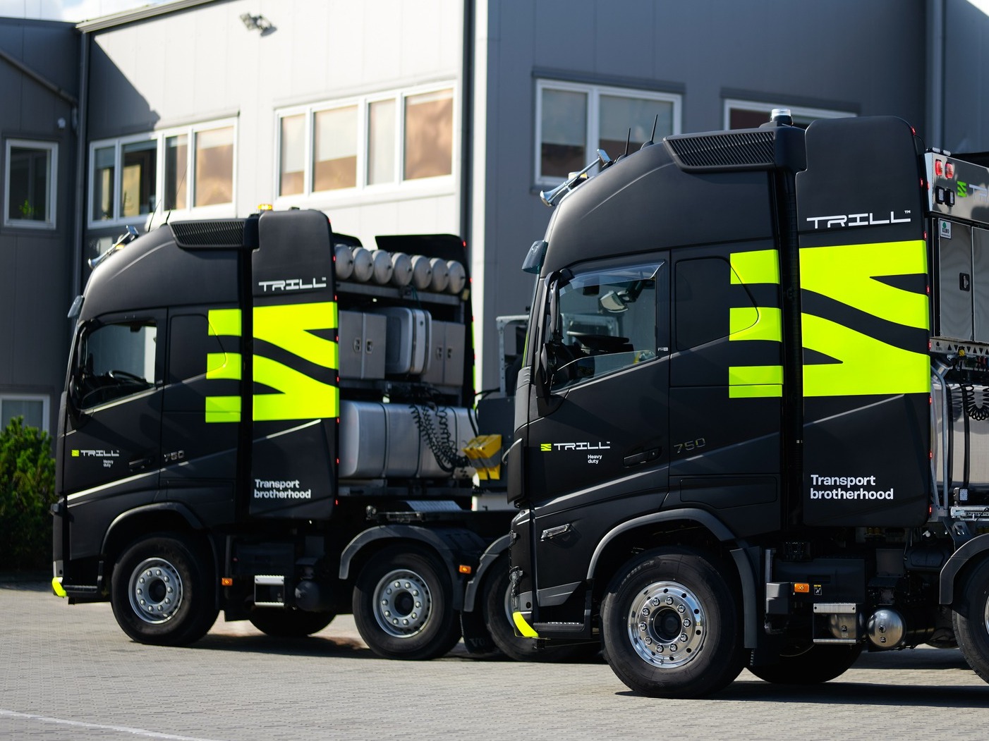 Dwa czarne ciągniki siodłowe z neonowo-zielonym logo 'TRILL' i napisem 'Transport brotherhood' na tle szarego budynku z oknami. Widok z boku, oświetlenie dzienne.