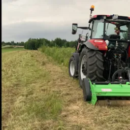 Czerwony traktor z zielonym agregatem koszącym na polu po koszeniu, widoczne ścięte źdźbła trawy i linia graniczna koszenia, pochmurne niebo w tle.