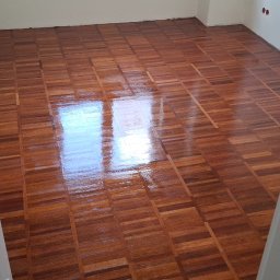 Lux-Parkiet - Błyszcząca, odnowiona podłoga parkietowa w jodełkę, widziana zza uchylonych białych drzwi. Parkiet po renowacji odbija światło okna. Ściany białe, widoczne gniazdka elektryczne.