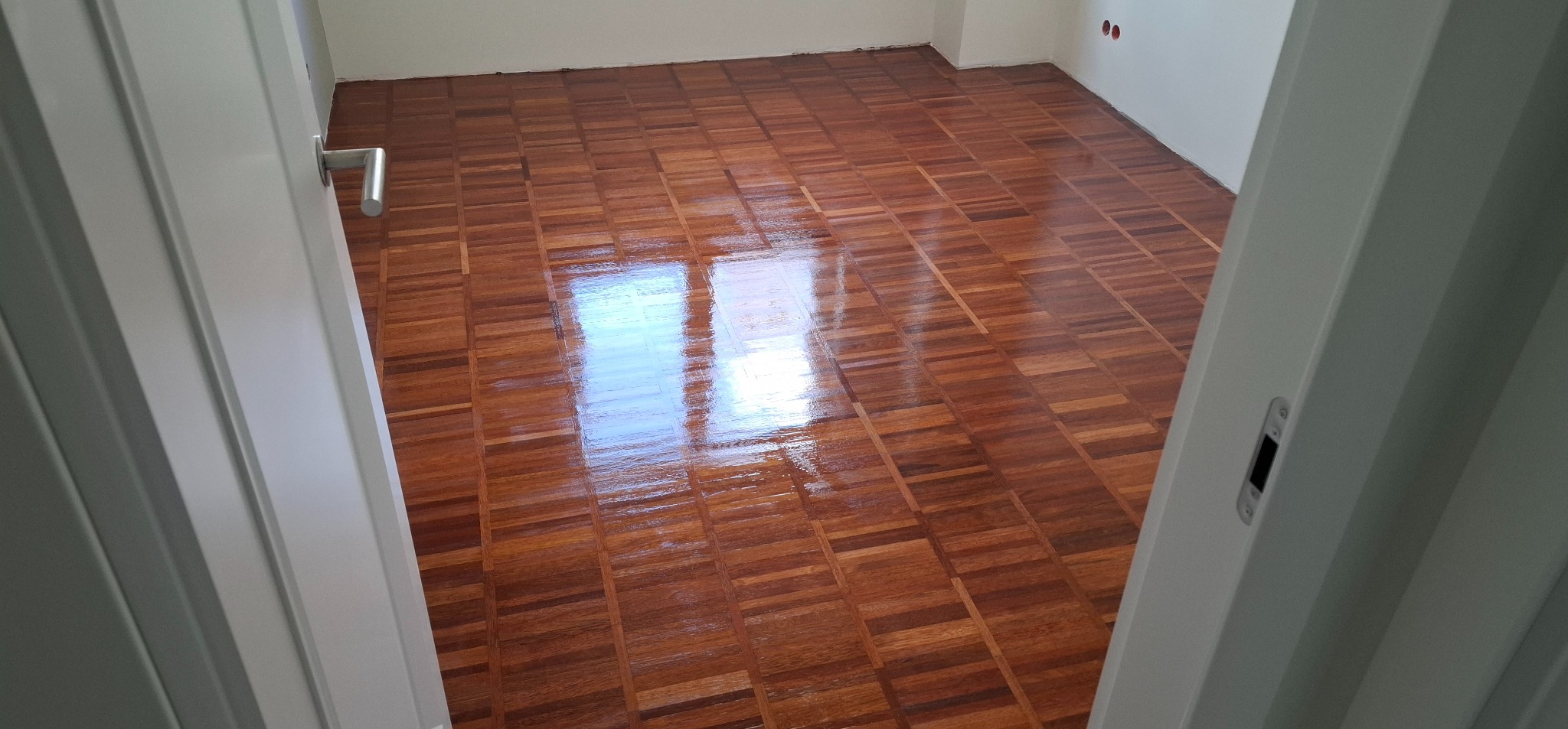 Błyszcząca, odnowiona podłoga parkietowa w jodełkę, widziana zza uchylonych białych drzwi. Parkiet po renowacji odbija światło okna. Ściany białe, widoczne gniazdka elektryczne.