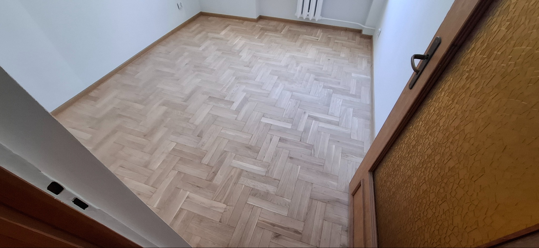 Parkiet ułożony w jodełkę francuską, widoczny z perspektywy wejścia do pokoju. Jasne drewno, białe ściany, fragment drzwi z ozdobnym szkłem.