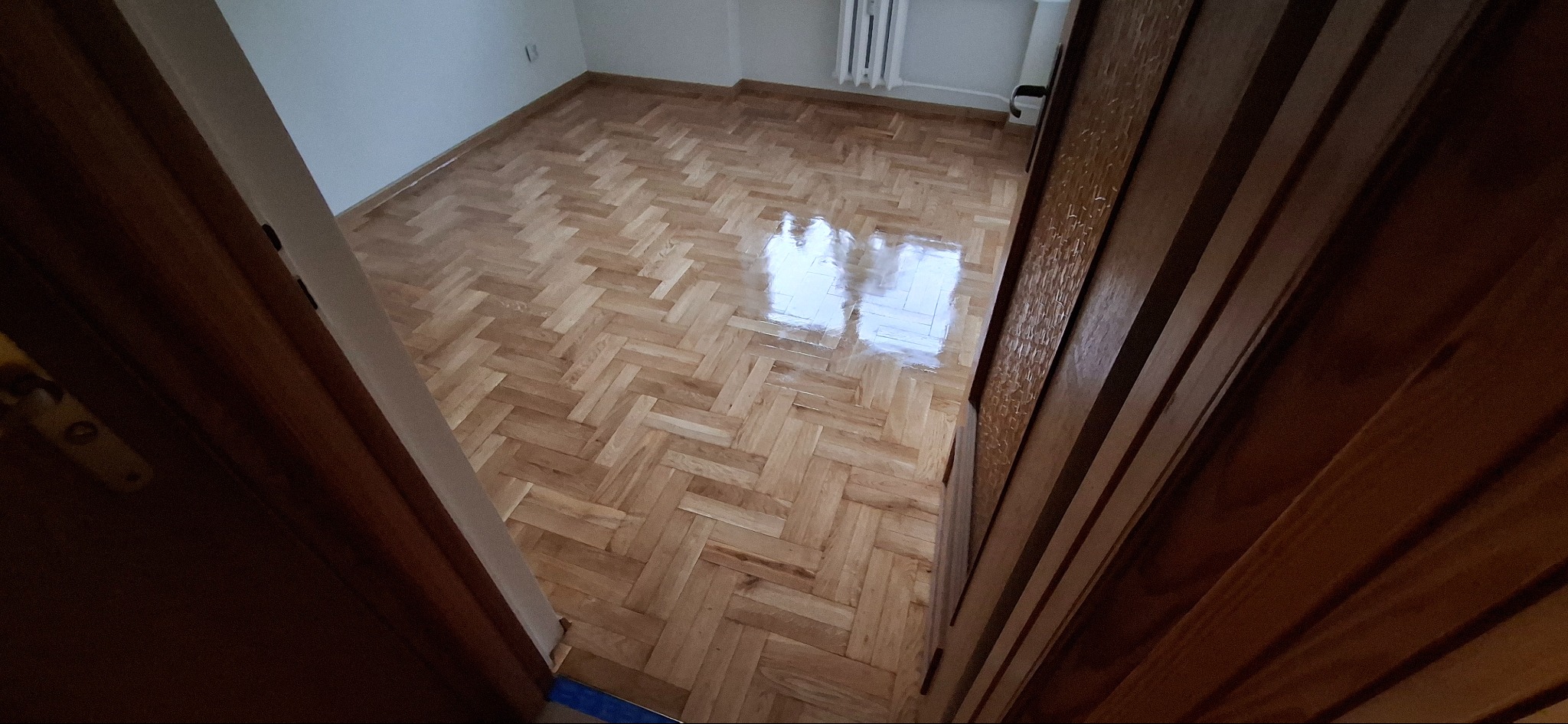 Błyszcząca, drewniana podłoga ułożona w jodełkę, widoczna zza uchylonych, ciemnych drzwi. Białe ściany i grzejnik w tle. Perspektywa z ukosa.