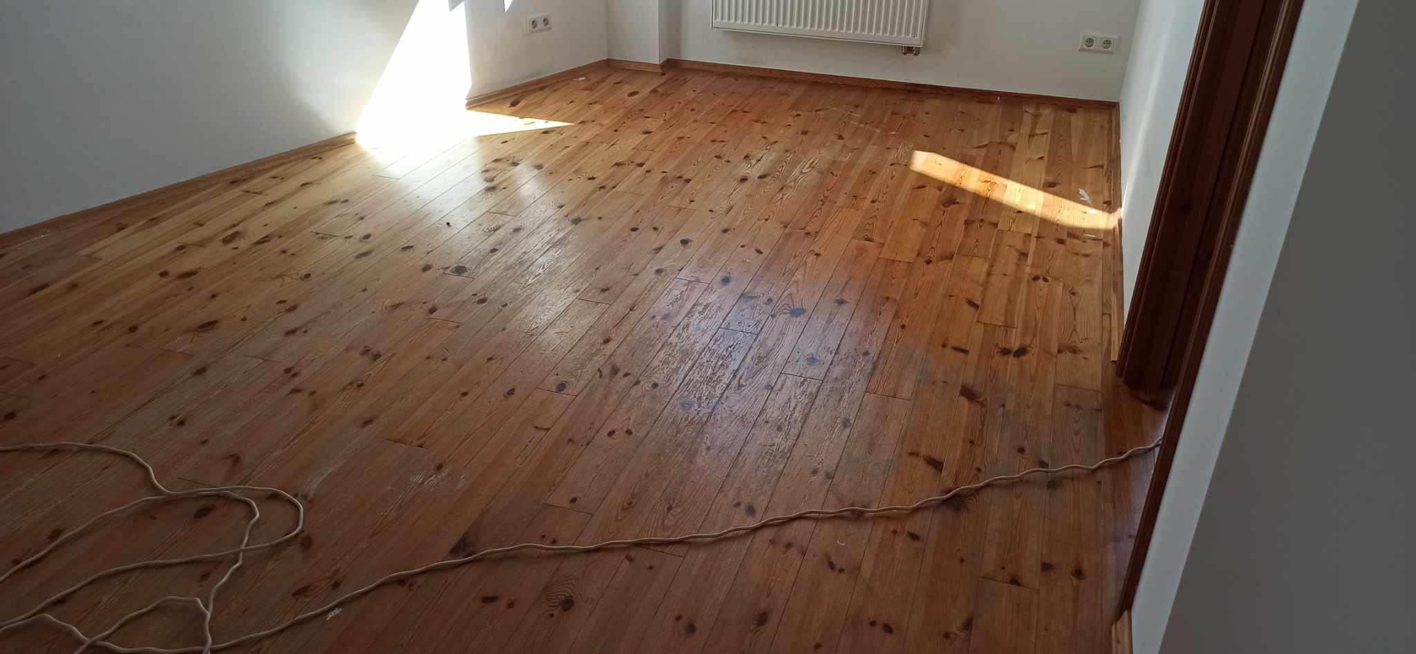 Wycyklinowana, lakierowana podłoga z desek w pustym pokoju, z widocznym przewodem elektrycznym i fragmentem grzejnika na ścianie.