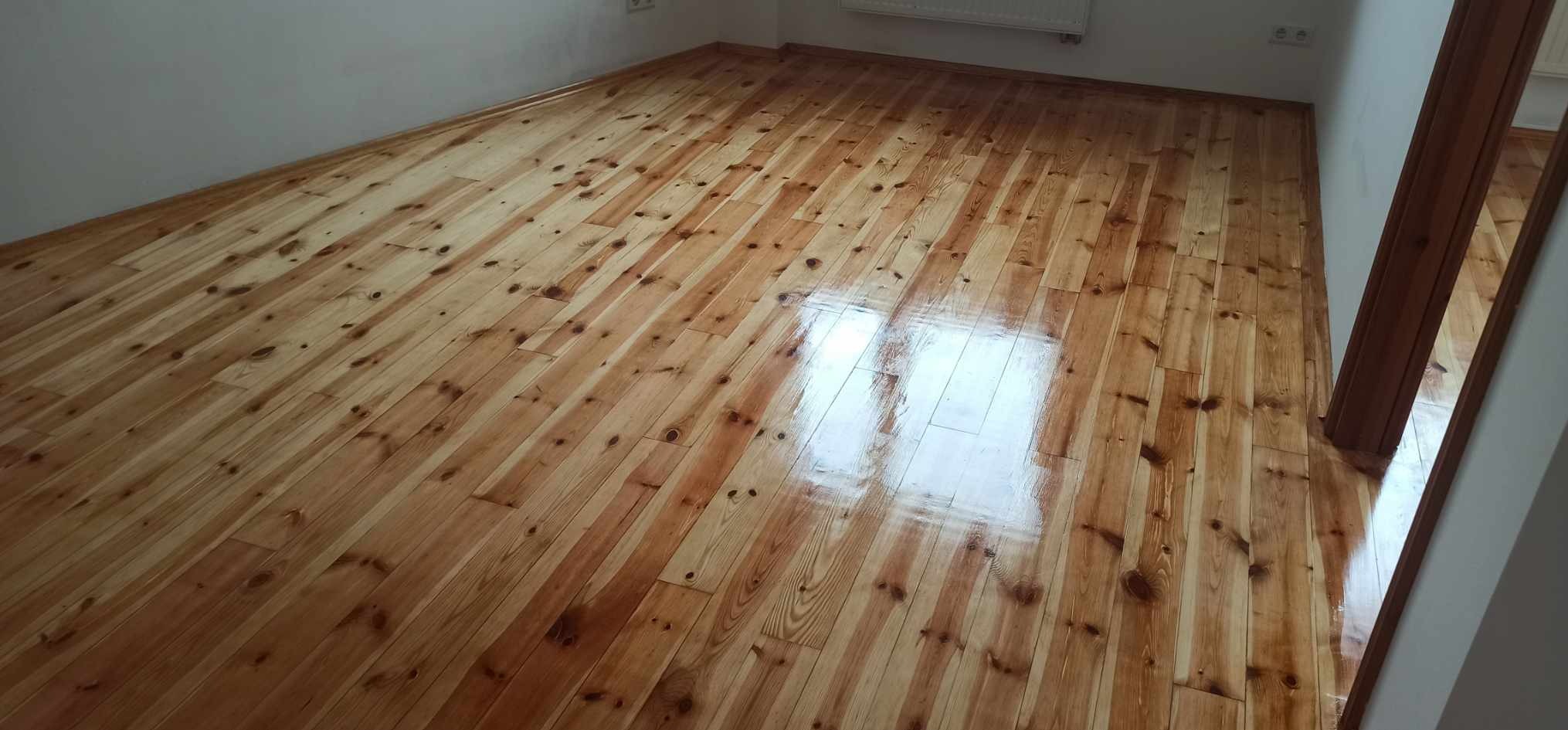 Błyszcząca, odnowiona drewniana podłoga z wyraźnym usłojeniem w jasnym pomieszczeniu. Widoczne ślady po cyklinowaniu i lakierowaniu, odbijające światło.