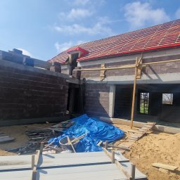 Usługi budowlane Kowalski - Budowa domu jednorodzinnego w trakcie realizacji: murowane ściany, więźba dachowa z łatami, niebieska plandeka, piasek. Widok na budynek z zewnątrz w słoneczny dzień.