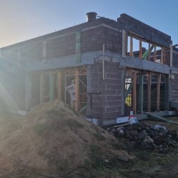 Usługi budowlane Kowalski - Budowa domu w trakcie realizacji: surowy stan z pustaków, widoczne drewniane szalunki okienne, w tle drzewa i zachodzące słońce. Na pierwszym planie sterta piasku.