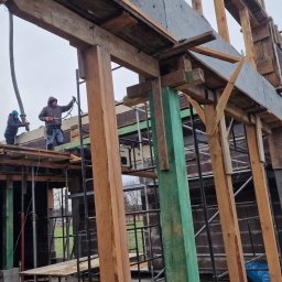 Usługi budowlane Kowalski - Budowa domu w toku: dwóch robotników na rusztowaniu pompuje beton do szalunków. Widoczne drewniane konstrukcje, bloczki i krajobraz wiejski w tle.