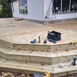 Usługi budowlane Kowalski - Drewniany taras z widocznymi schodami i narzędziami, częściowo pokryty opadłymi liśćmi. W tle dom z białej cegły i drabina. Widok z poziomu gruntu.