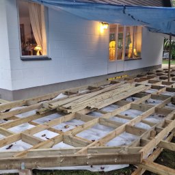 Usługi budowlane Kowalski - Konstrukcja drewniana tarasu przy białym domu z oknami, zabezpieczona folią. Widoczne elementy podtrzymujące i izolacja pod konstrukcją. Prace w toku, oświetlenie w oknach.