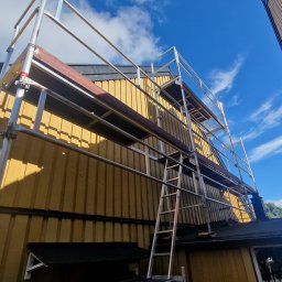 Usługi budowlane Kowalski - Żółta elewacja domu w trakcie prac na wysokościach, widoczna z dołu z fragmentem rusztowania i drabiny na tle błękitnego nieba.