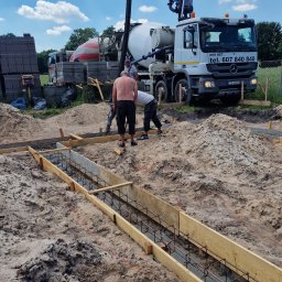 Usługi budowlane Kowalski - Wylewanie fundamentów pod budynek za pomocą betonomieszarki z pompą. Widoczne deskowanie, zbrojenie i pracownicy. W tle zaparkowany samochód ciężarowy.