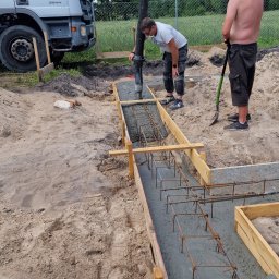 Usługi budowlane Kowalski - Wylewanie betonu do szalunku zbrojeniowego pod fundament budynku. Pracownicy obsługują pompę do betonu, widoczny samochód betoniarki i teren budowy.