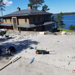 Układanie kostki brukowej na posesji w Grudziądzu. Widoczny fragment ułożonej nawierzchni, pracownicy i narzędzia. W tle dom i jezioro.